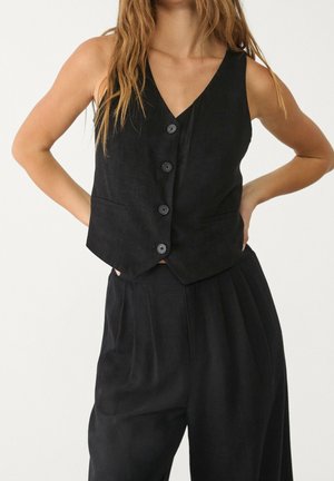 Woman wearing sleeveless black buttoned vest and loose black pants, standing with hands on hips against white background.