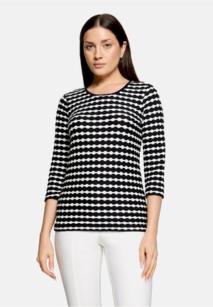 Femme aux cheveux raides et foncés portant un haut à manches trois-quarts noir et blanc à motifs et un pantalon blanc, debout devant un fond uni.