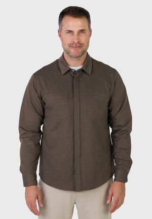 Homme portant une chemise à manches longues marron avec des poches poitrine sur un t-shirt blanc, debout devant un fond uni.