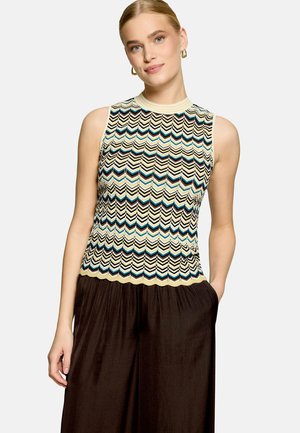 Femme aux cheveux blonds portant un top en tricot sans manches à motif chevrons crème, noir, bleu et marron, associé à un pantalon large marron foncé.