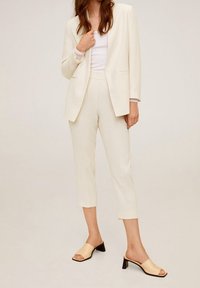 Cremefarbener Blazer und passende verkürzte Hose, kombiniert mit beigen offenen Mules mit schwarzem Blockabsatz. Glattes Material mit taillierter Passform.
