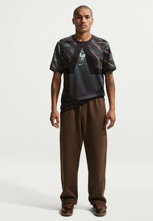 Άνδρας όρθιος, φοράει μαύρη αθλητική φανέλα με σχέδια και λογότυπο κόκορα, καθώς και καφέ φαρδιά παντελόνια, με ορατά τατουάζ στα μπράτσα.