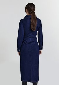 Femme avec une longue queue de cheval brune, portant une longue robe en denim bleu foncé et des bottes noires, de dos contre un fond clair.