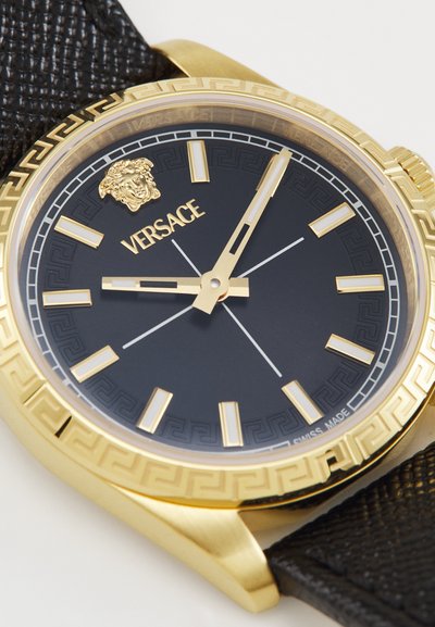 Kuldtooni käekell musta dialsiga, paks kuldne raami, millel on Kreeka võtme muster, ja tekstureeritud mustast nahast rihm. Versace logo on eksponeeritud.