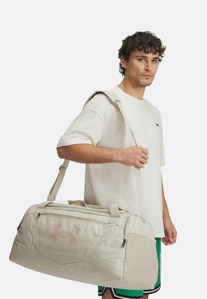 Beige reistas met ritsen, bovenhandvatten en een schouderriem. Heeft het Under Armour-logo en textuurvakken van stof. Duurzaam materiaal.
