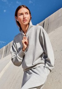 Grijze sweatshirt met een hoge kraag en een half-rits ontwerp, gemaakt van zacht, gestructureerd stof. Draagt in combinatie met bijpassende broek, tegen een blauwe lucht.