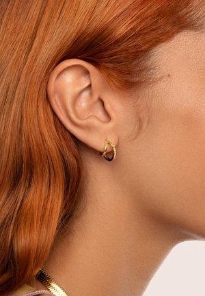 Golden hoop-Ohrringe mit einer gedrehten Textur, präsentiert vor einem Hintergrund aus glatter Haut und rotem Haar, wobei ihre reflektierende Oberfläche zur Geltung kommt.