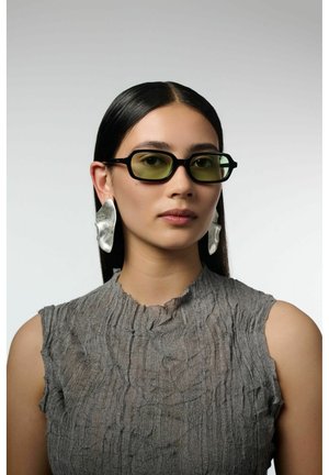 Femme aux cheveux noirs raides portant des lunettes rectangulaires teintées de vert, de grandes boucles d'oreilles argentées abstraites, et un haut gris texturé sans manches.