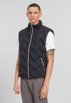 Schwarweste aus gepolstertem Material mit hohem Kragen, Reißverschluss und zwei Fronttaschen. Getragen über einem weißen T-Shirt und kombiniert mit grauen Hosen. Glatte Textur.