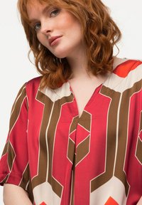 Kurzarmpullover mit V-Ausschnitt in Rot und Braun, mit geometrischen Mustern und kontrastierenden cremefarbenen Akzenten. Glatte, leichte Stoffqualität.