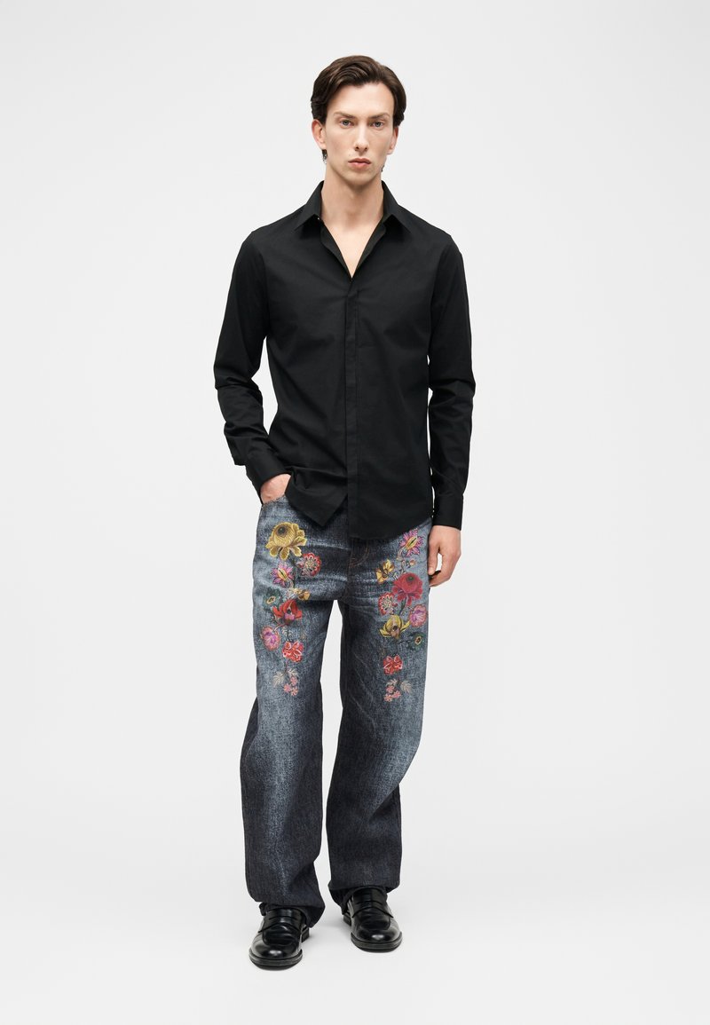 Homme portant une chemise noire à boutons et un jean ample sombre orné de broderies florales colorées, debout avec une main dans la poche.