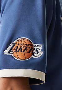 Blue sports jersey featuring an embroidered “Los Angeles Lakers” logo with a basketball. White trim accents the sleeve.