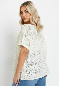 Top de punto blanco de gran tamaño con un corte holgado, que presenta patrones de calados y mangas cortas dobladas. Combinado con jeans de mezclilla azules.