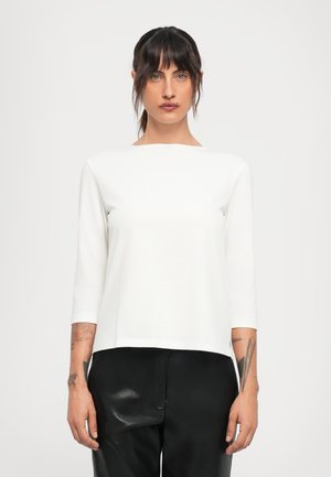 Junge Frau mit dunklem Haar und Tattoos trägt ein weißes Langarmshirt und schwarze Lederhosen und steht vor einem schlichten hellen Hintergrund.