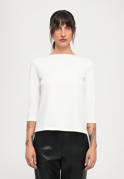 Jeune femme aux cheveux foncés et tatouages portant une chemise blanche à manches longues et un pantalon en cuir noir, se tenant devant un fond léger uni.