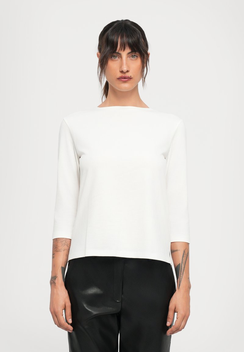 Young woman with dark hair and tattoos wears white long-sleeve shirt and black leather pants, standing against a plain light background.