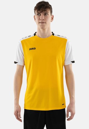 Jonge man die een geel sportshirt met korte witte mouwen en zwarte korte broek draagt, staande tegen een effen achtergrond.