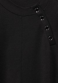 Haut en tissu noir avec un tricot texturé, un col rond et une patte de boutonnage avec cinq boutons noirs brillants sur l'épaule gauche.