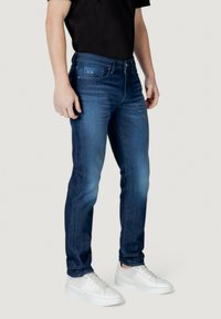 Vaqueros de mezclilla de color azul oscuro en un corte slim, con un diseño deslavado, estilo de cinco bolsillos y un sutil logo en la cinturilla. Combinados con zapatillas blancas.