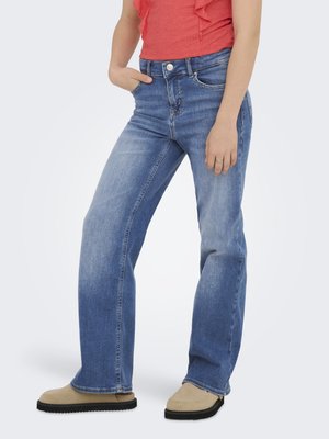 Moteris dėvi vidutinio plovimo tiesaus kirpimo džinsus, koralinės spalvos rankovių neturintį viršutinį drabužį ir smėlio spalvos slip-on batus, stovėdama su viena ranka kišenėje.