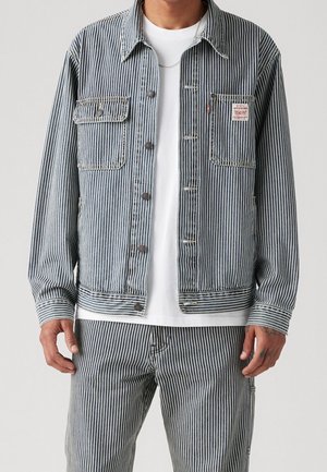 Homme portant une veste rayée verticale bleue et blanche et un pantalon assorti sur une chemise blanche, montrant les détails avant et des poches.