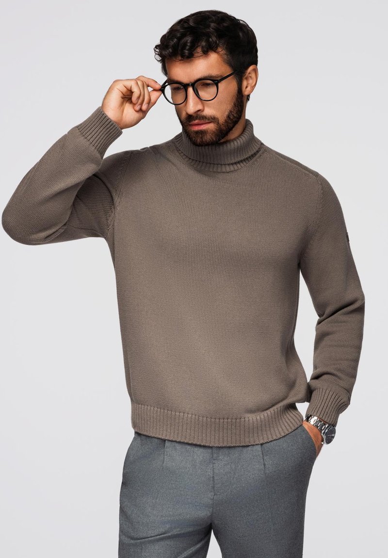 Brązowy sweter z golfem w prążki, z długimi rękawami, wykończonymi mankietami i dołem; noszony z szarymi spodniami; model dostosowuje okulary.