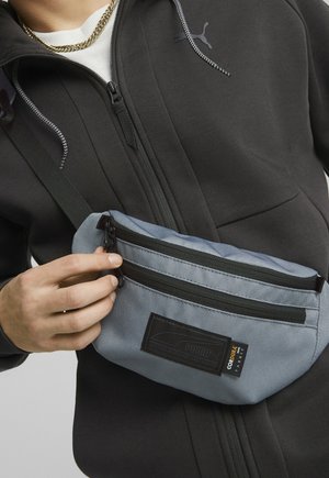 Main en train d'ajuster la fermeture éclair d'un sac banane Puma bleu clair, porté sur une veste zippée noire avec le logo Puma et un collier en chaîne dorée.