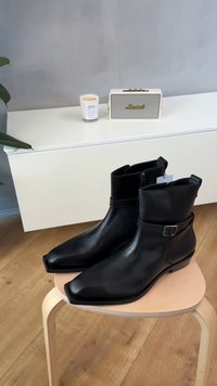 Μαύρα δερμάτινα μποτάκια αστραγάλου με μυτερή μύτη, κλείσιμο με φερμουάρ και διακοσμητικό λουράκι με αγκράφα στον αστράγαλο, τοποθετημένα σε ένα ξύλινο σκαμπό.