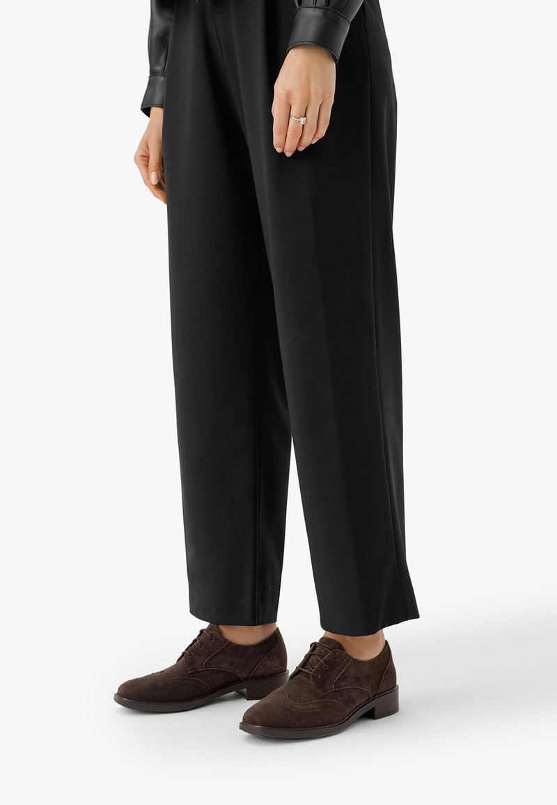 Persona che indossa pantaloni neri a gamba larga e scarpe in suede marrone con lacci, con una mano visibile che mostra un anello a taglio quadrato.