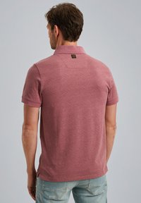 Polo à manches courtes de couleur rose, avec un motif texturé. Il dispose d'un col classique et d'une étiquette à l'arrière, associé à un jean bleu clair.