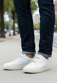 Białe wsuwane trampki z teksturowaną skórzaną cholewk�ą, beżowymi elastycznymi wstawkami i gumową podeszwą, zestawione z podwiniętymi ciemnymi dżinsami.