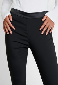 Schwarze Leggings aus einem glatten, dehnbaren Stoff mit breitem Satinbund. Das Design verfügt über subtile Seitennähte und eine körperbetonte Silhouette.