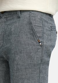 Graue Hosen im Fischgrätmuster mit strukturiertem Finish, ausgestattet mit seitlichen Taschen und braunen Hardwareakzenten.