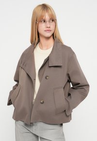 Femme blonde portant des lunettes transparentes, un pull beige, une grande veste marron à deux boutons et un pantalon gris clair, les mains dans les poches de la veste.