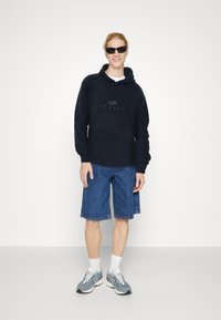 Felpa navy con tasca frontale, abbinata a pantaloni shorts in denim. Le calzature sono scarpe da ginnastica grigie con calze bianche a crew.