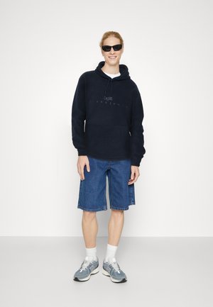 Haut à capuche bleu marine avec une poche avant, associé à un short en denim. Les chaussures sont des baskets grises avec des chaussettes blanches.