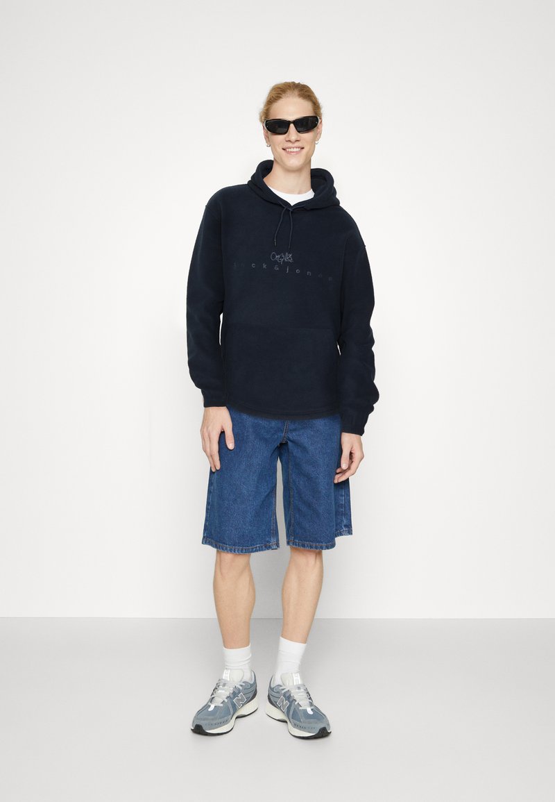 Felpa navy con tasca frontale, abbinata a pantaloni shorts in denim. Le calzature sono scarpe da ginnastica grigie con calze bianche a crew.