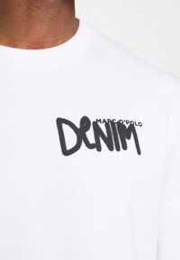 Vit sweatshirt med en framträdande svart broderad logotyp som säger "DENIM" och "MARC O'POLO" i mindre text ovanför.