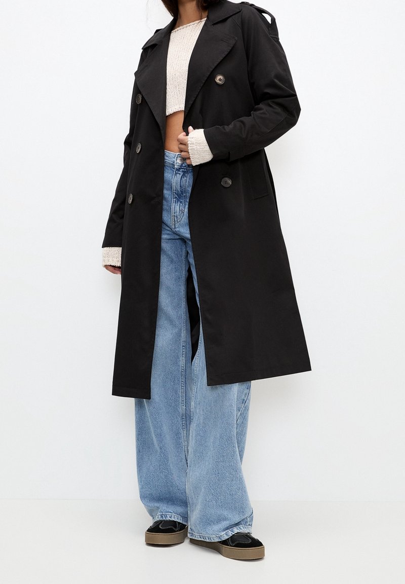 Femme portant un long trench-coat noir par-dessus un pull court beige, un jean large bleu et des baskets noires à plateforme sur un fond blanc uni.