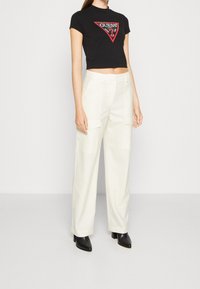 Schwarzes, kurzes T-Shirt mit rotem und weißem "GUESS"-Logo, kombiniert mit cremefarbenen, weiten Cargo-Hosen. Das Model trägt schwarze High-Heels.