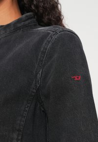 Μαύρο τζιν μπουφάν με δομημένο γιακά, που διαθέτει κόκκινο κεντημένο λογότυπο στο μανίκι. Το κοντινό πλάνο δείχνει την υφή του υφάσματος και τις ραφές.