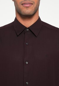 Bordó gombos ing klasszikus gallérral, sima anyagból és fekete gombokkal. Bal oldalon kis CK logó található.