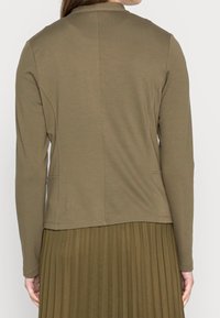 Olive green long-sleeve top with a round neckline, smooth fabric, and side pockets, paired with a pleated, olive green skirt.