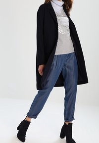 Lungo cappotto nero sopra una felpa grigia con stampa, abbinato a jeans blu chiaro con risvolto e stivali neri alla caviglia. Tessuto morbido e vestibilità rilassata.