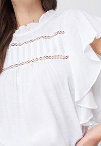 Blouse blanche en tissu léger, avec des manches volantes, un corps texturé et une bande décorative avec des découpes.