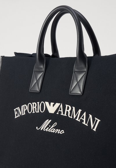 Borsa tote in tela nera con manici in pelle nera, caratterizzata da testo ricamato bianco "EMPORIO ARMANI Milano" in un design curvo.