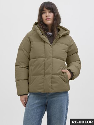 Kvinde iført en olivengrøn dunjakke med hætte og blå jeans, stående med den ene hånd i lommen mod en ensfarvet lys baggrund.