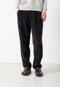 Muškarac u crnim manšestrastim hlačama, sivom džemperu i crnim cipelicama stoji nasuprot bijeloj pozadini.