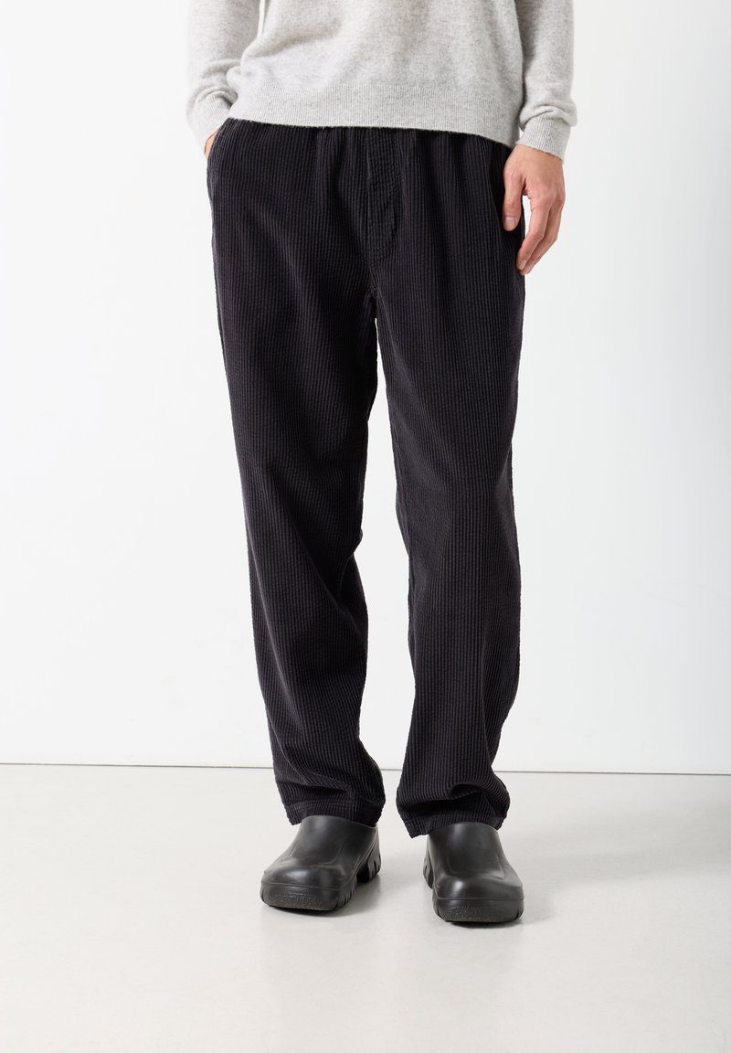 Muškarac u crnim manšestrastim hlačama, sivom džemperu i crnim cipelicama stoji nasuprot bijeloj pozadini.