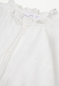 White linen garment featuring a ruffled neckline and a partial front zip. Soft texture with a subtle striped pattern. Label visible.